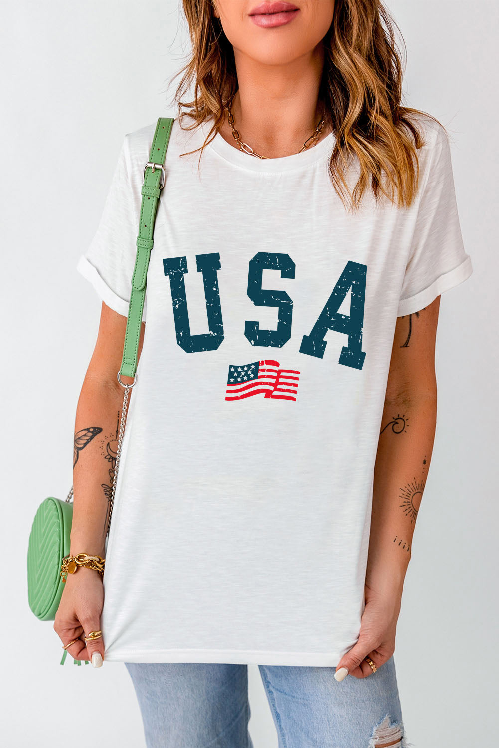 Stylish woman models a casual white USA print t-shirt with distressed jeans and a trendy green crossbody bag, evoking a relaxed patriotic vibe. Perfect for subtle national pride.