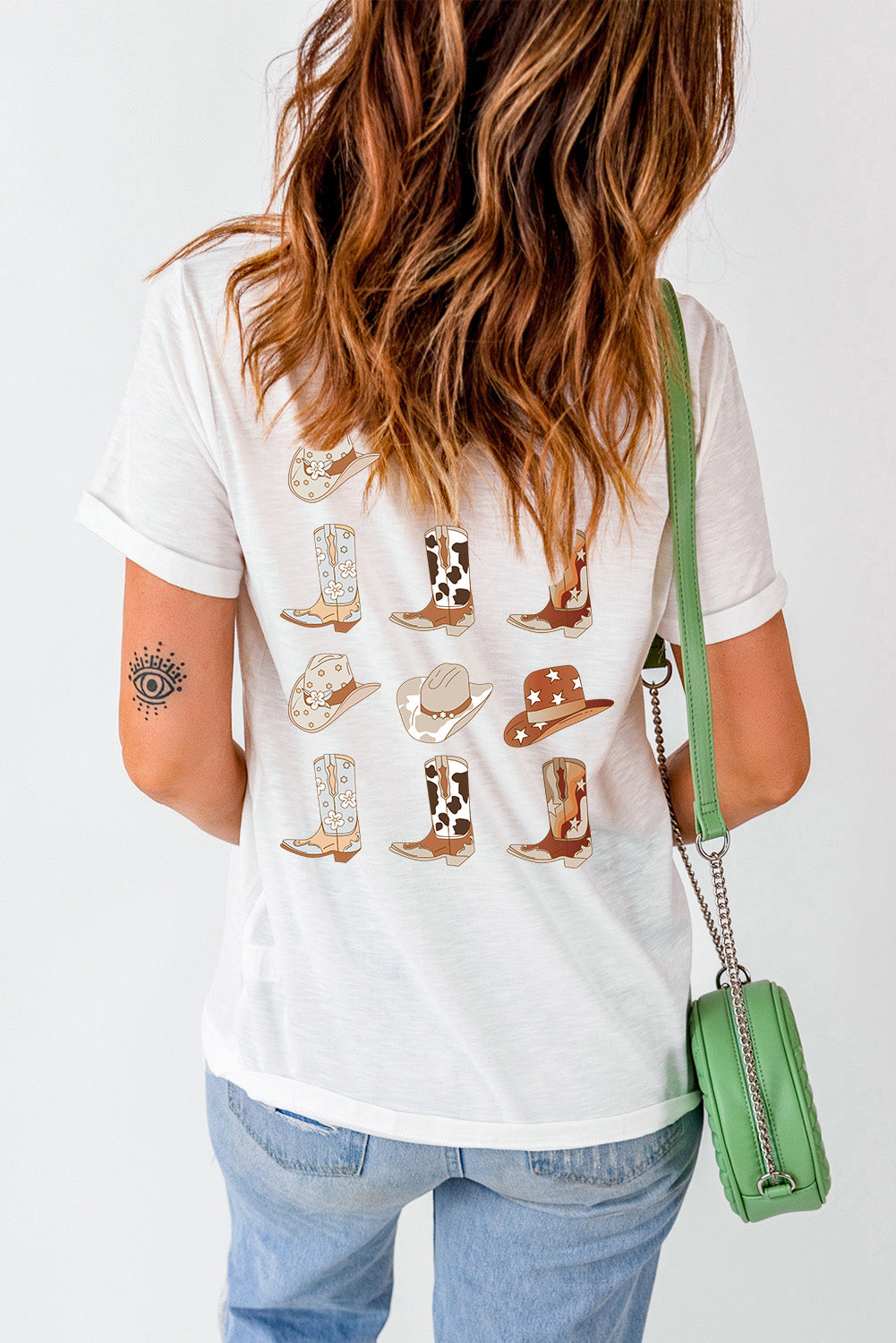 Woman in a white T-shirt with cowboy boots and hat prints, blue jeans, carrying a green shoulder bag, showcasing casual style.