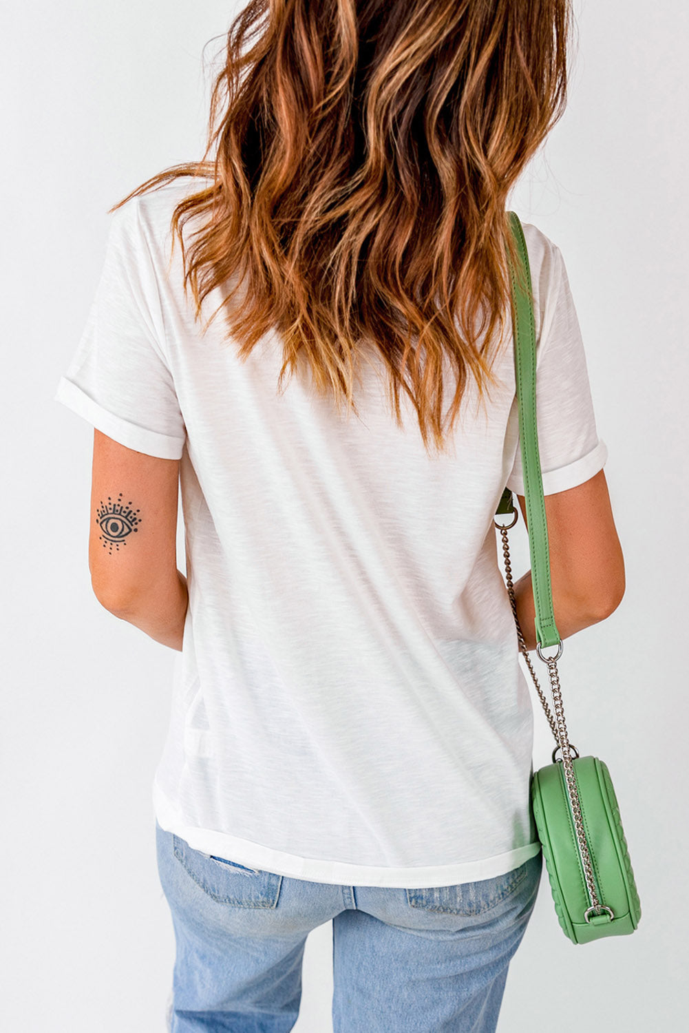 Woman in white tee and blue jeans sporting an eye tattoo, holding a chic green chain-strap bag. Perfect for casual style.