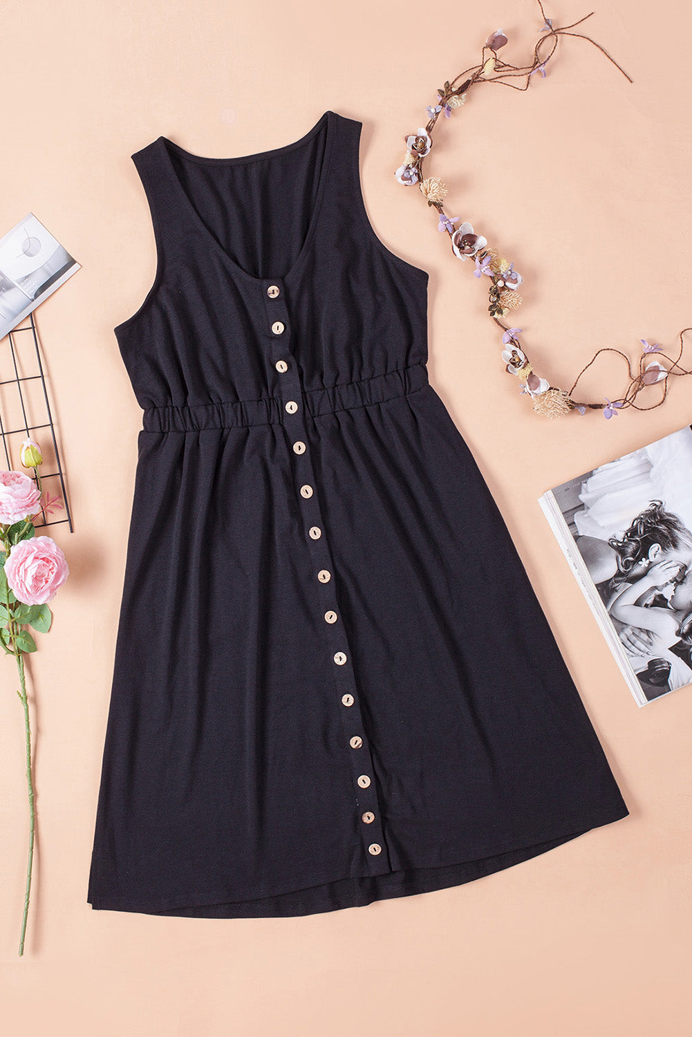 Black sleeveless dress with button details and a cinched waist, displayed flat lay with floral accents on a peach background. Perfect for a casual summer look.