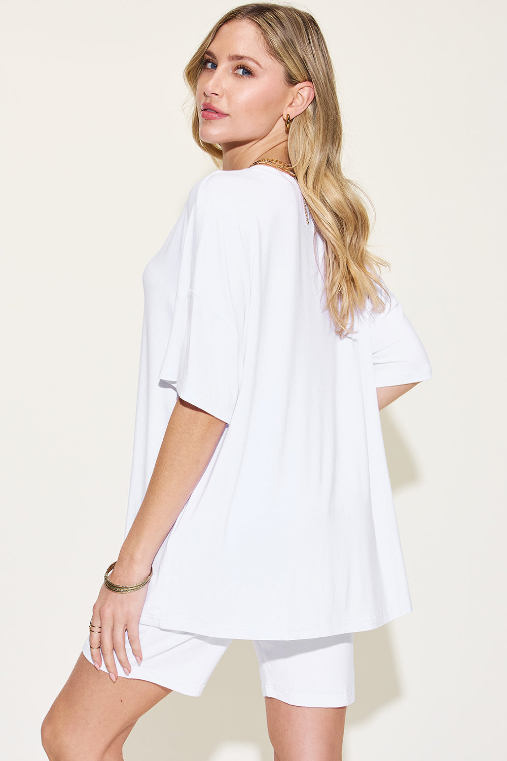 Elegant woman in a white oversized t-shirt and shorts, showcasing casual summer fashion with a subtle pose, on a cream background.