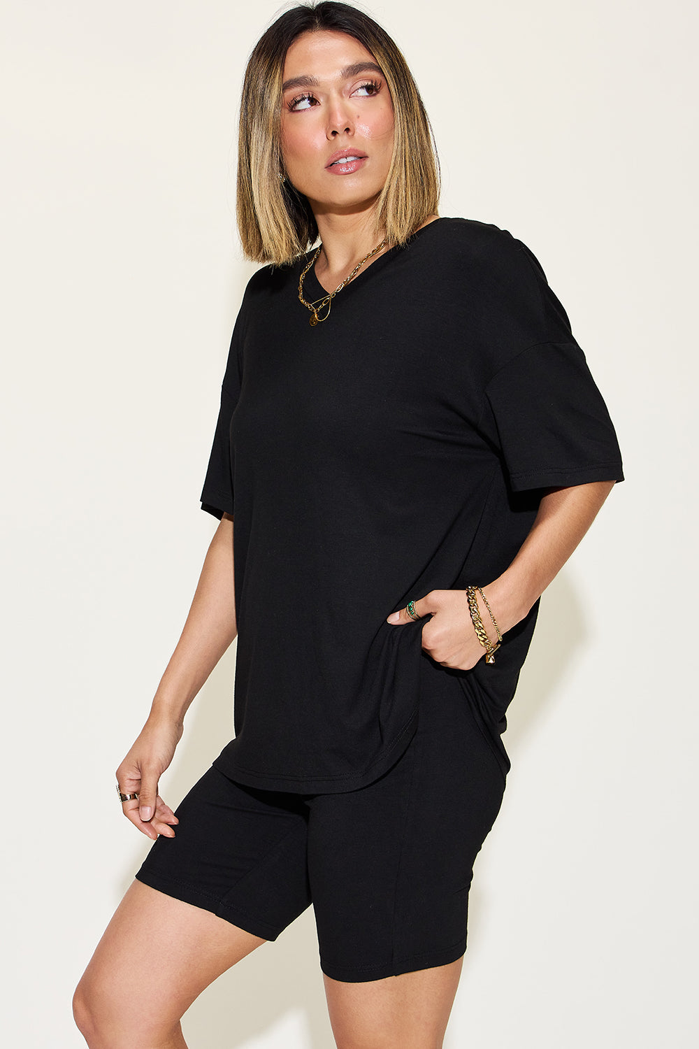 Chic woman modeling a versatile black t-shirt and matching shorts, complemented by gold-tone jewelry, against a neutral backdrop—perfect for a casual summer outfit.