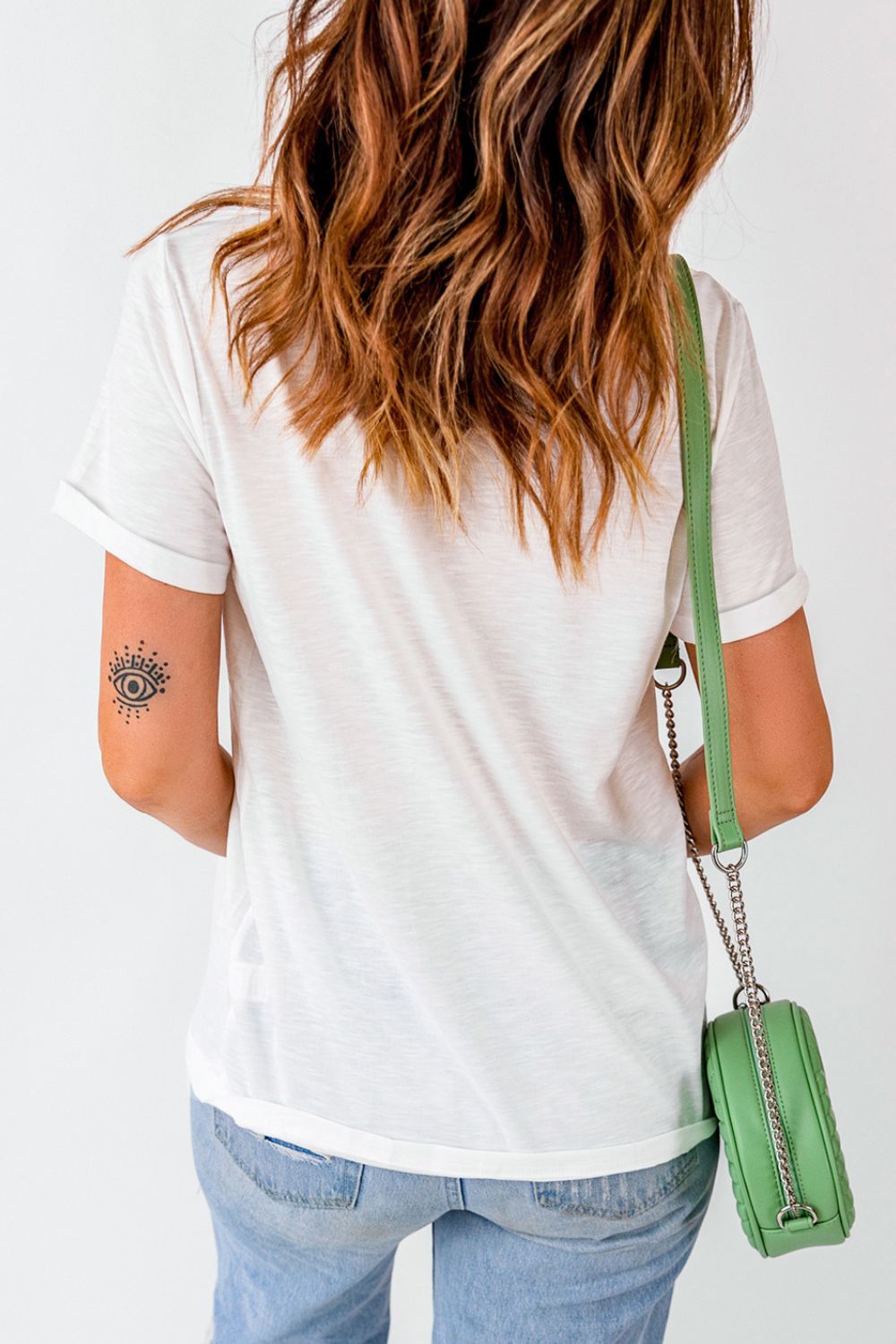 Woman in casual white tee and blue jeans carrying a stylish green shoulder bag with a metallic chain. Visible small arm tattoo adds a trendy touch to the effortless chic look. Perfect for fashion and lifestyle content.