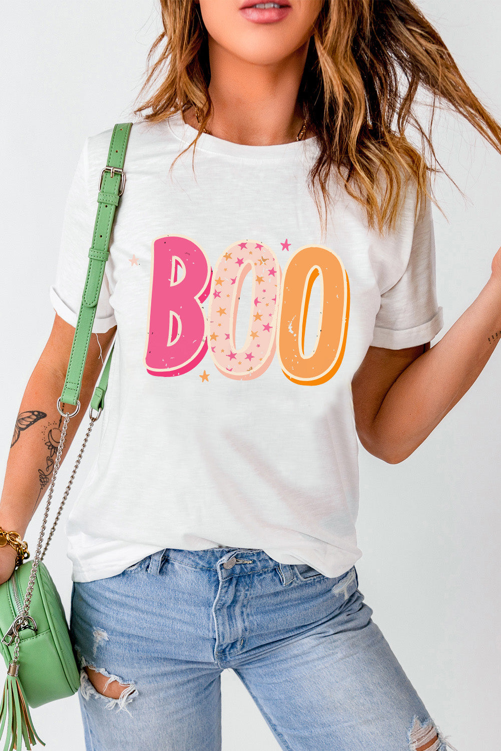 Woman in casual white tee with festive 'BOO' print paired with distressed jeans and a green crossbody bag, embodying a chic yet relaxed style perfect for a laid-back outing.