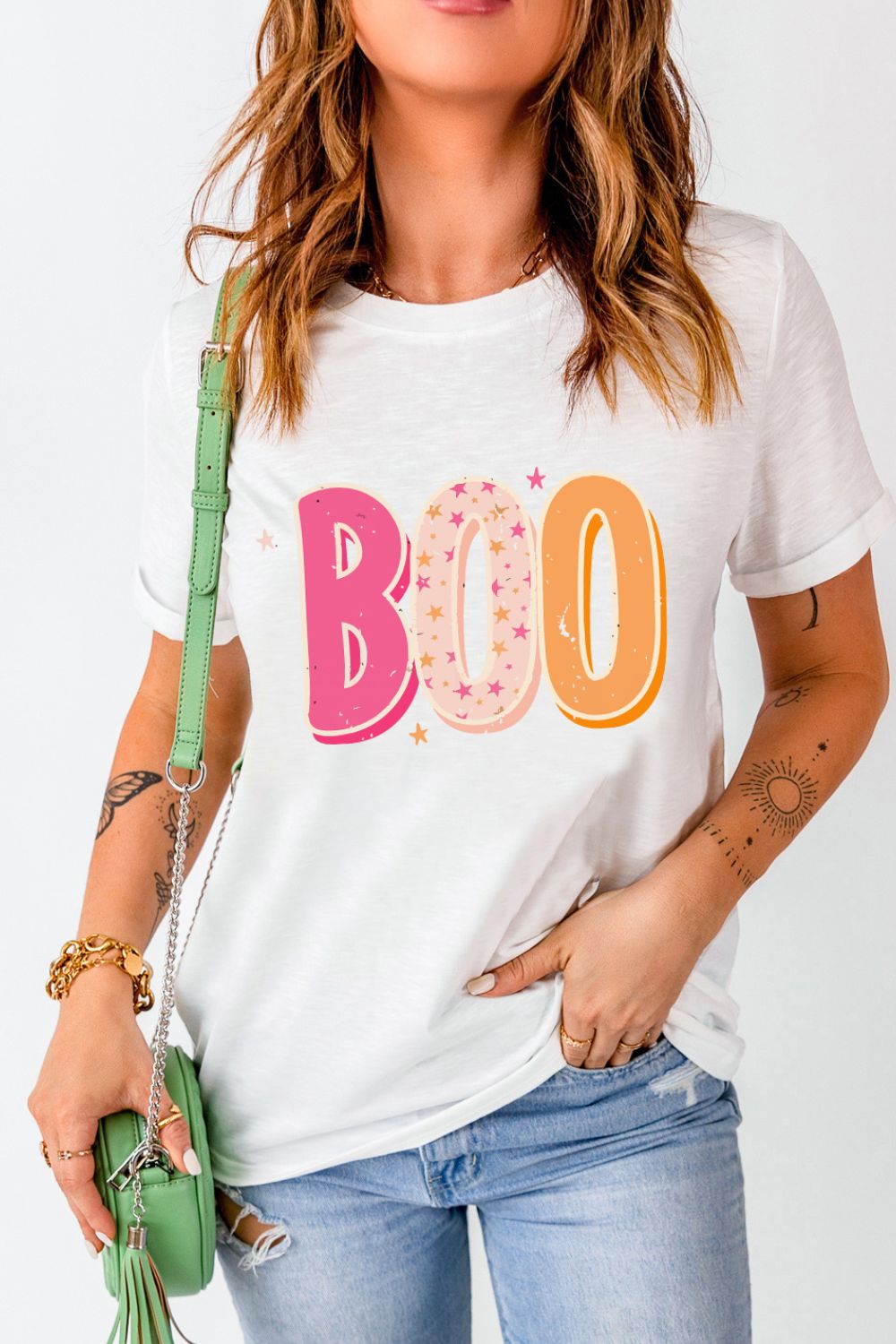 Woman in casual style with a graphic "BOO" tee, carrying a trendy green crossbody bag, and stylish accessories, perfect for a laid-back look.