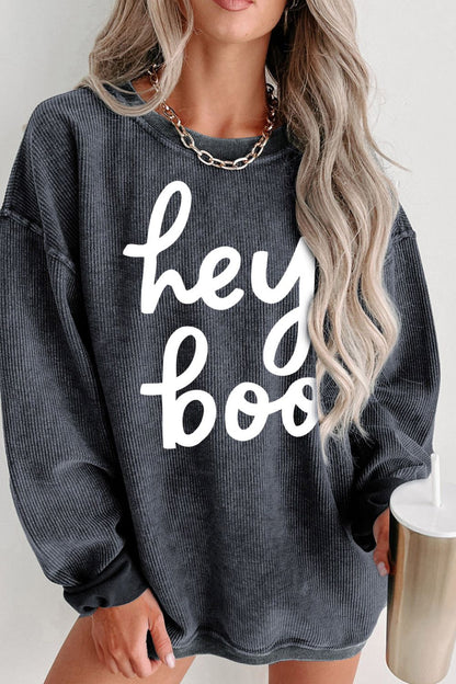 Casual charcoal gray oversized sweatshirt with white "hey boo" text, featuring a stylish woman with wavy blonde hair accessorized with a chunky necklace, holding a trendy metallic tumbler—a perfect autumn fashion statement.