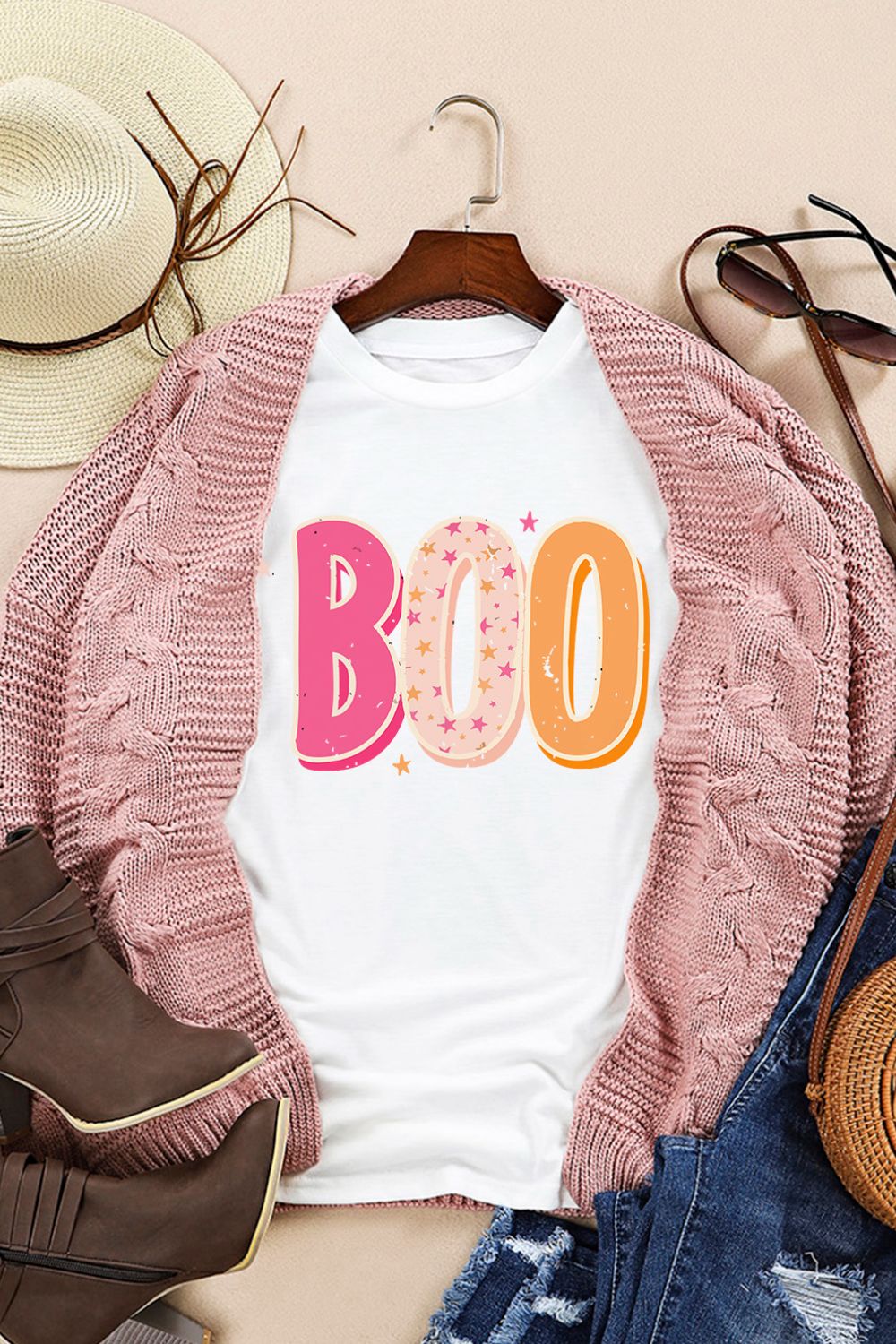 Stylish autumn outfit with a playful "BOO" graphic tee, pink knit cardigan, distressed jeans, ankle boots, sun hat, and sunglasses for a casual chic look. Perfect for fall fashion and Halloween vibes.