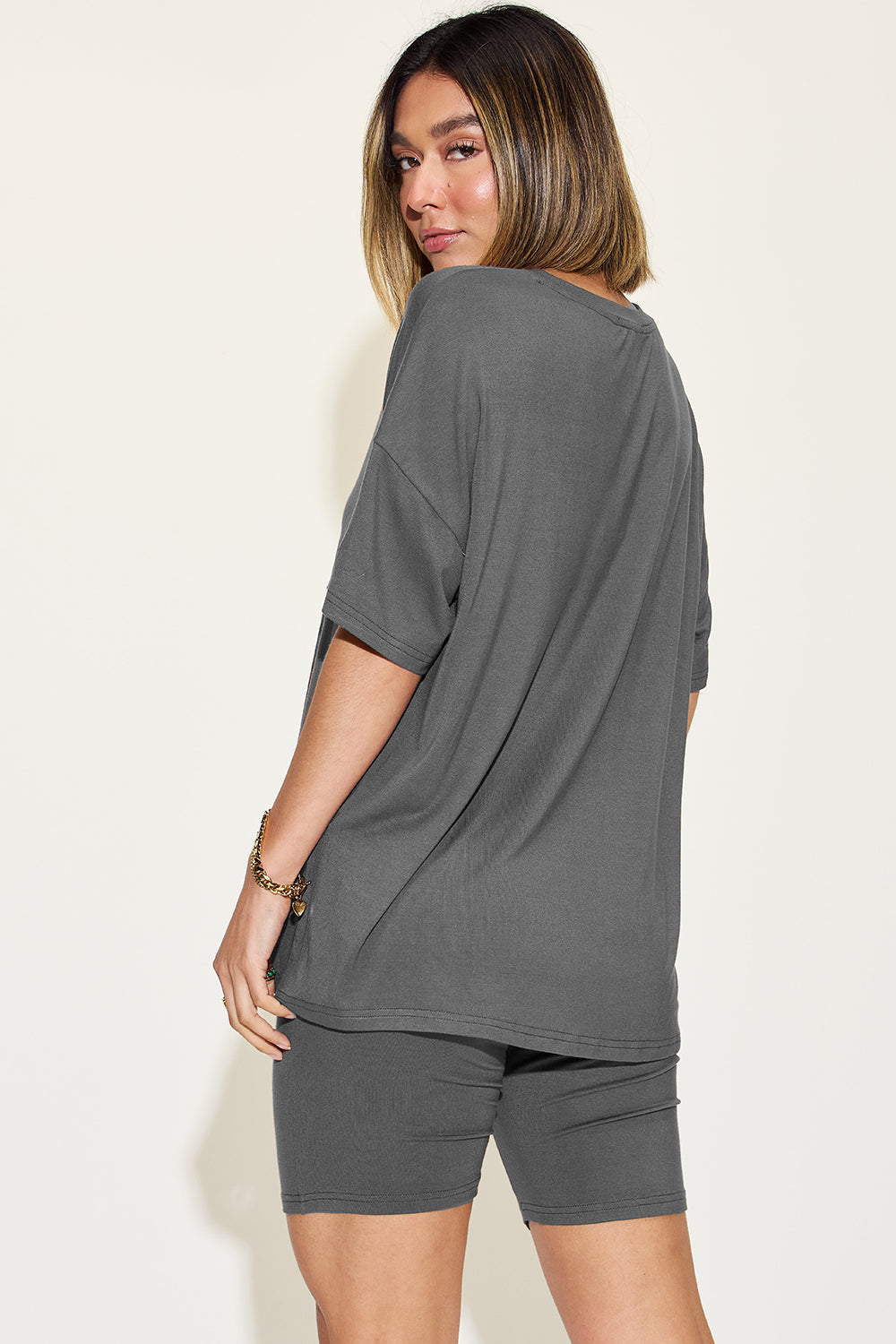 Woman in a gray oversized t-shirt and cycling shorts glancing back, sporting a casual summer look with subtle jewelry. Perfect for a relaxed, stylish outfit.