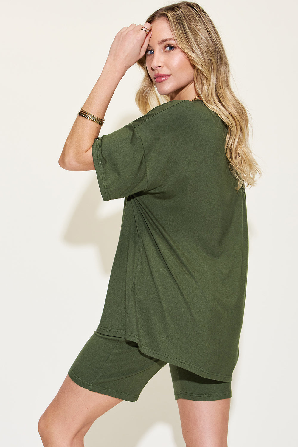 Blonde woman in casual olive green t-shirt dress, posing with hand on head, showcasing relaxed streetwear style and gold bracelets.