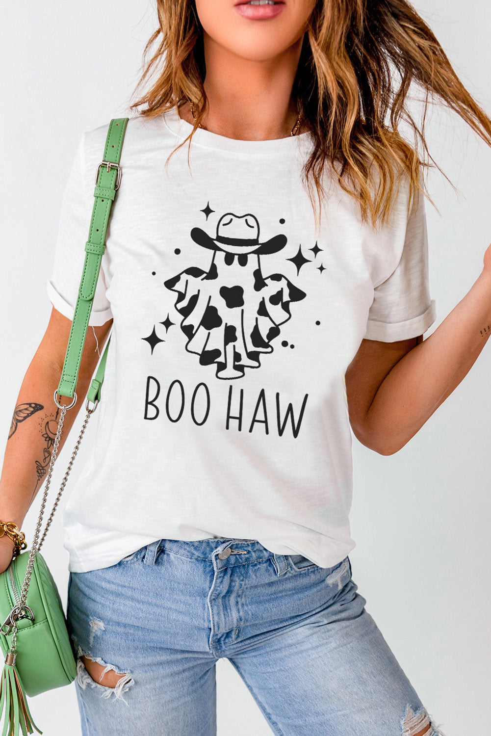 Woman in white t-shirt with "BOO HAW" text and playful ghost in cowboy hat illustration, paired with distressed blue jeans and green shoulder bag.